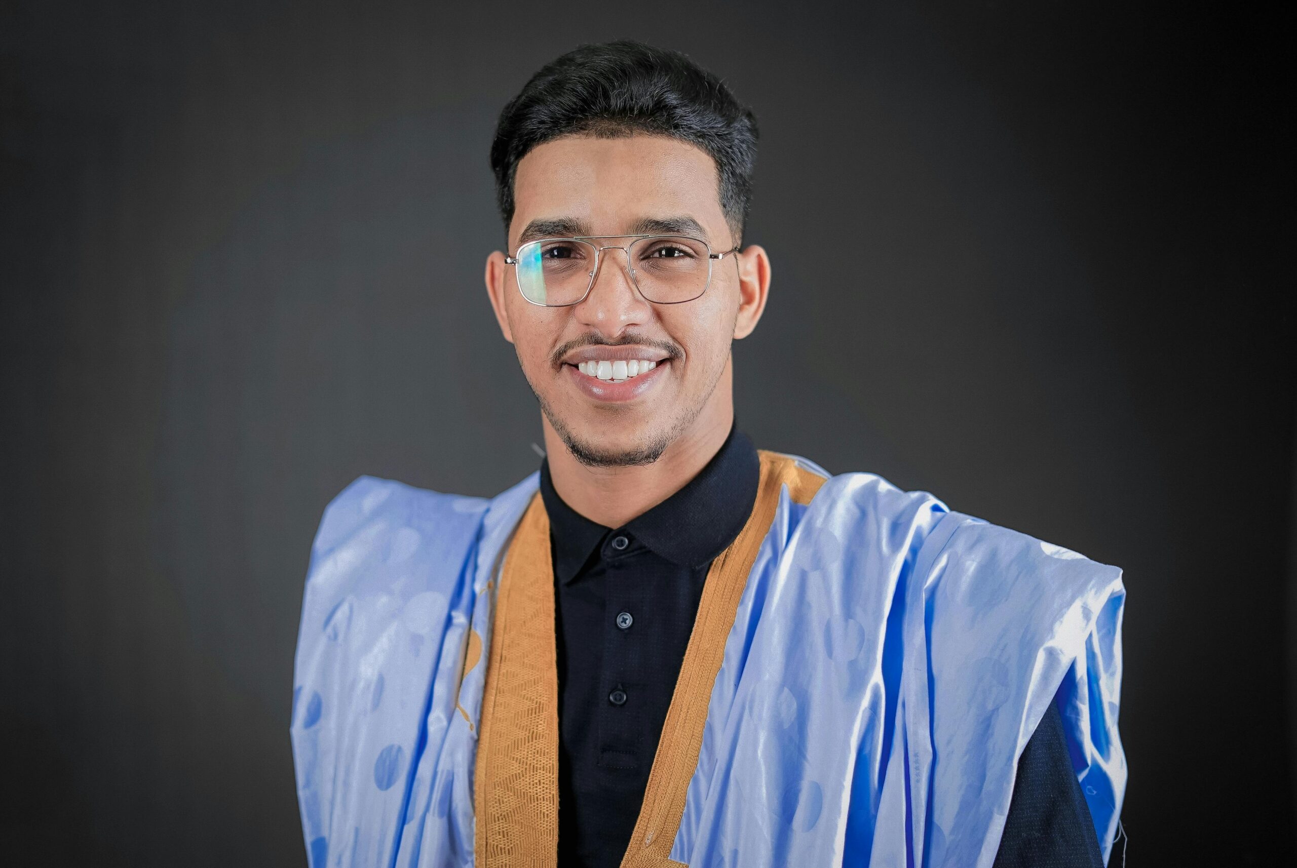 Portrait of a smiling man in traditional clothing with eyeglasses against a gray background.